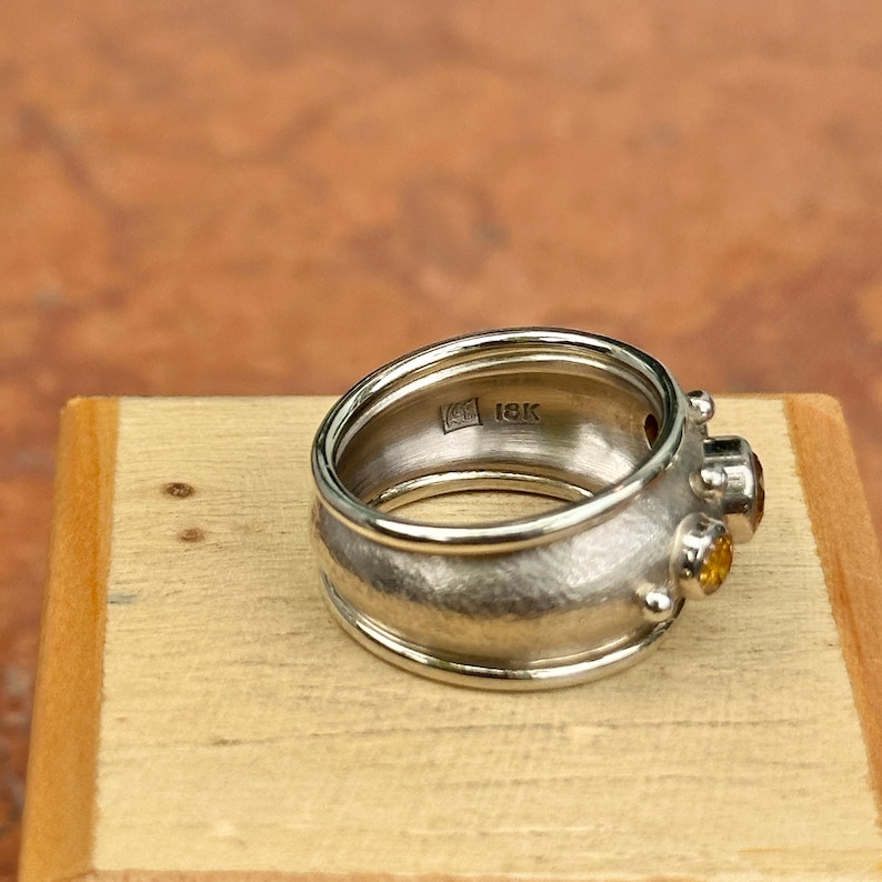 May include: A silver ring with a textured band and two small, round, yellow gemstones. The ring has an 18K stamp inside the band. The ring is displayed on a wooden surface.