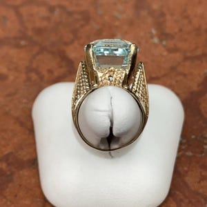 May include: A gold-toned ring featuring a large, rectangular, light blue gemstone. The ring has an ornate, textured design on the band and around the gemstone setting. The ring is displayed on a white stand against a brown background.