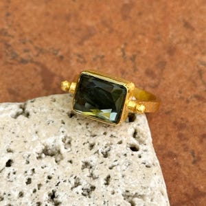 May include: A gold-toned ring with a rectangular, dark green gemstone. The ring has a textured band and small decorative elements on either side of the gemstone. The ring is displayed on a light-colored, textured surface.