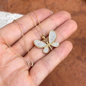 May include: A gold-colored butterfly pendant necklace with a delicate chain. The butterfly pendant is encrusted with small, sparkling white stones, and the wings have a detailed, textured design. The necklace is displayed against a neutral background.
