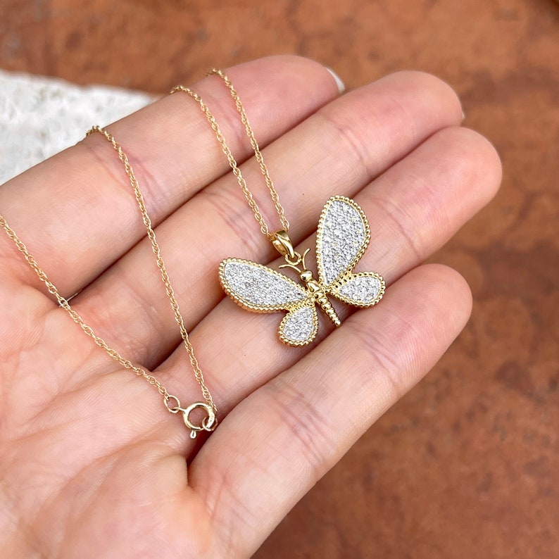May include: A gold-colored butterfly pendant necklace with a delicate chain. The butterfly pendant is encrusted with small, sparkling white stones, and the wings have a detailed, textured design. The necklace is displayed against a neutral background.