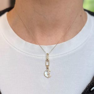 May include: A gold-colored necklace with a crescent moon pendant. The pendant features a mother-of-pearl inlay and a star accent. The necklace is worn on a white shirt.