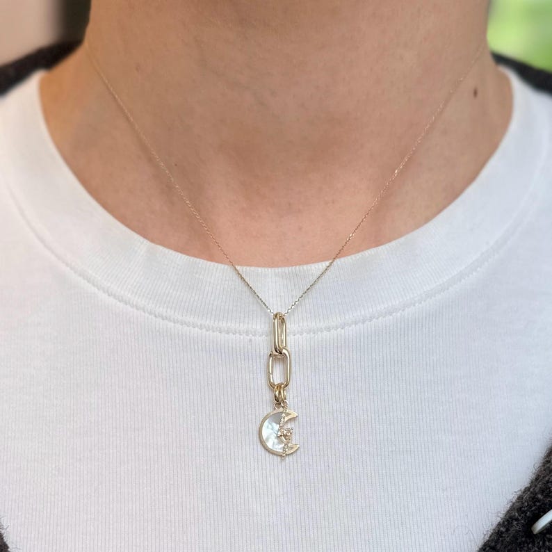 May include: A gold-colored necklace with a crescent moon pendant. The pendant features a mother-of-pearl inlay and a star accent. The necklace is worn on a white shirt.