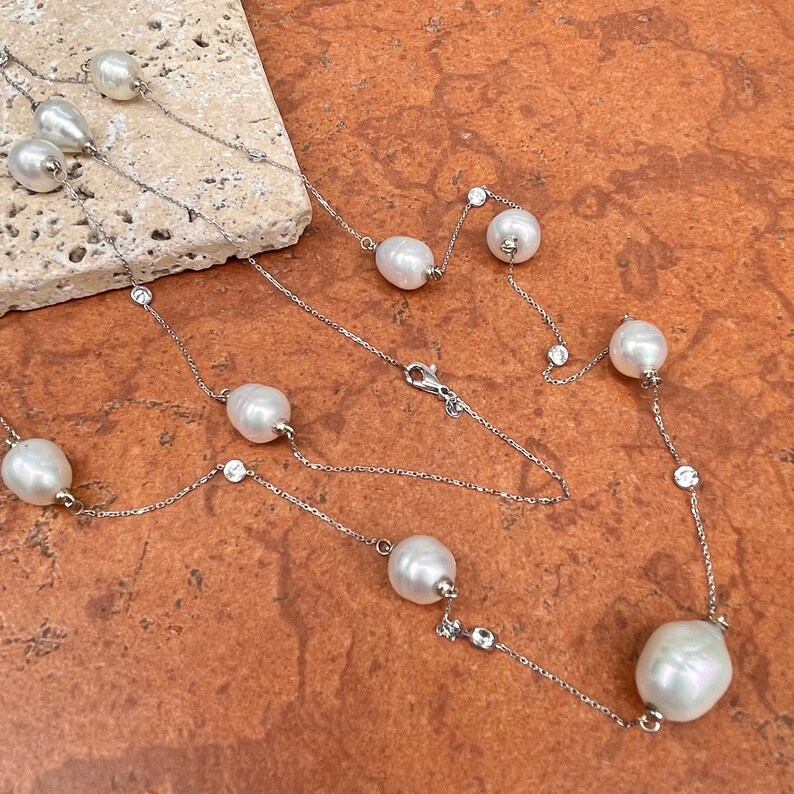 K&ouml;nnte beinhalten: Eine zarte Halskette mit gl&auml;nzenden wei&szlig;en Perlen und kleinen, funkelnden, klaren Kristallen. Die Perlen sind unregelm&auml;&szlig;ig geformt und entlang einer feinen Silberkette angeordnet. Die Halskette wird auf einer strukturierten, hellfarbigen Oberfl&auml;che vor einem warmen, r&ouml;tlich-braunen Hintergrund pr&auml;sentiert.
