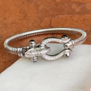 May include: A silver-toned metal bracelet featuring a flexible, ribbed band and a central horseshoe-shaped clasp. The clasp is embellished with small, clear pavé-set stones and two dark, round cabochon accents at the connection points.