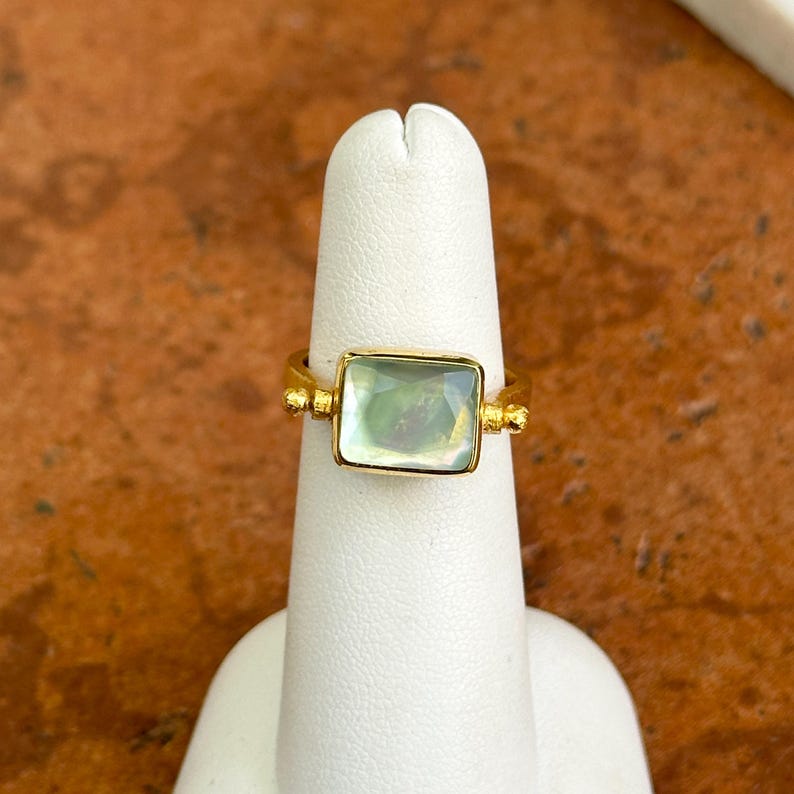 May include: A gold-toned ring with a square, iridescent gemstone. The ring has decorative details on the band and around the gemstone setting. The gemstone appears to shift colors in the light, creating a unique look. This ring is a piece of jewelry.