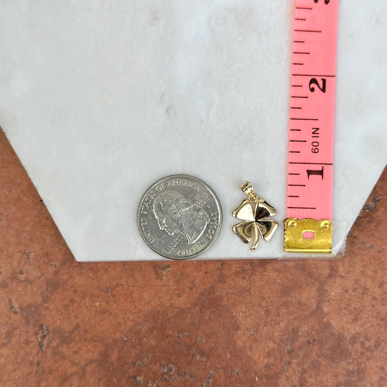 May include: A gold four-leaf clover charm pendant, measuring approximately 2.5 cm in length, is shown next to a US quarter for size comparison.