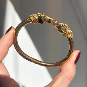 May include: A gold-colored bracelet featuring two horse head sculptures facing each other. The horse heads have green gemstone eyes. The bracelet is held by a person's hand.