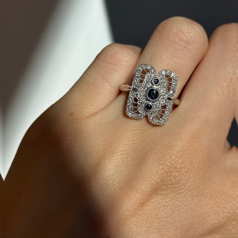 May include: A silver ring with a geometric design featuring three blue gemstones surrounded by smaller white gemstones.