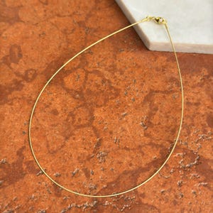 May include: A delicate gold-coloured necklace with a thin, curved design. The necklace has a simple clasp and is displayed on a textured, reddish-brown surface with a white marble accent.