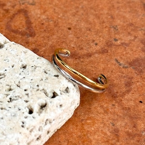May include: A two-tone open ring featuring a polished gold-colored band layered over a silver-colored band. The ring has a smooth, rounded finish and an open design, allowing for adjustment. The metal bands are joined at the ends.