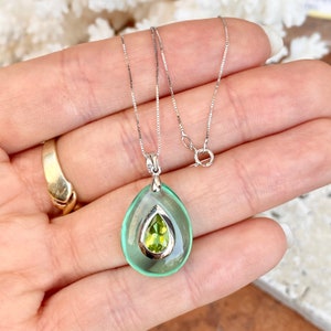 May include: A silver necklace with a teardrop-shaped pendant. The pendant has a light green outer layer and a smaller, pear-shaped, light green gemstone in the center. The necklace is displayed on a hand.