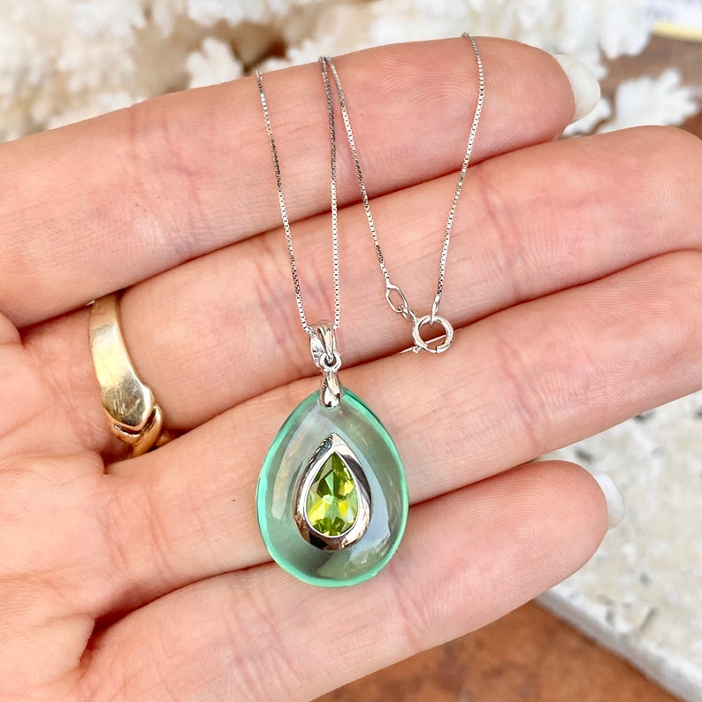 May include: A silver necklace with a teardrop-shaped pendant. The pendant has a light green outer layer and a smaller, pear-shaped, light green gemstone in the center. The necklace is displayed on a hand.