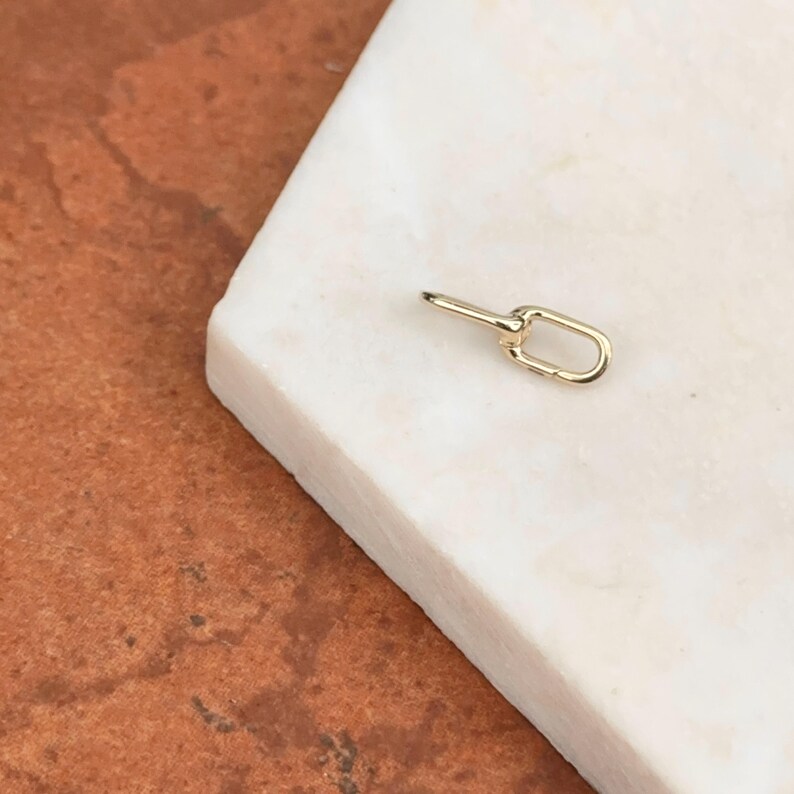 May include: A small, gold-colored jewelry clasp with an oval loop and a straight bar. The clasp is lying on a white marble surface, with a warm-toned, reddish-brown background.