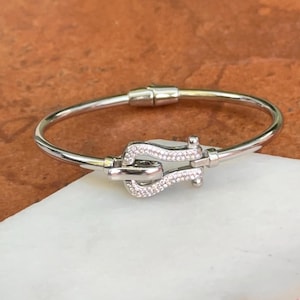 May include: A silver-toned metal bangle bracelet featuring a central horseshoe-shaped clasp. The clasp is embellished with numerous small, clear pavé-set stones that cover the entire surface of the horseshoe element, creating a sparkling effect.