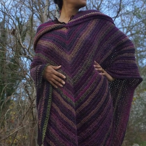 Puede incluir: Un chal de crochet a rayas de color morado, verde y marr&oacute;n con capucha. El chal est&aacute; drapeado sobre los hombros de una persona.