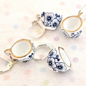 Pode incluir: Brincos de cerâmica branca em forma de chávenas de chá em miniatura, com desenhos florais azuis e detalhes dourados. Os brincos têm fechos de orelha prateados. As chávenas de chá são detalhadas, mostrando uma estética delicada e feminina.