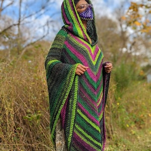 Puede incluir: Un poncho de crochet con capucha en tonos de verde, negro y morado. El poncho tiene un patr&oacute;n de chevron y lo lleva puesto una persona con una m&aacute;scara facial morada.