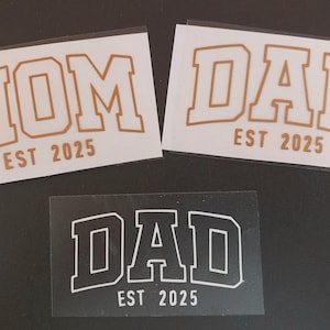 以下が含まれることがあります： 黒い輪郭のある白いアイロン接着ビニールステッカー3枚。ステッカーには「MOM EST 2025」、「DAD EST 2025」、「DAD EST 2025」と書いてあります。