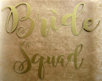 May include: Gold foil letters spelling out "Bride Squad" on a brown paper background.