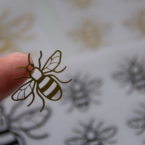 May include: A gold bee sticker with black stripes on a white background. The sticker is being held by a finger.