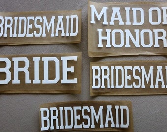 May include: Five white iron-on patches with the words "BRIDESMAID", "BRIDE", "MAID OF HONOR", "BRIDESMAID", and "BRIDESMAID" on a brown background.