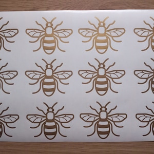 May include: Sheet of gold-coloured bee stickers. Each sticker features a detailed outline of a bee with wings and striped abdomen. The stickers are arranged in a grid pattern on a white background.
