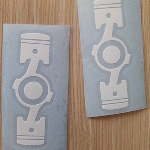 May include: Two gray stickers with a white outline of a piston. The piston is a common symbol used in the automotive industry.