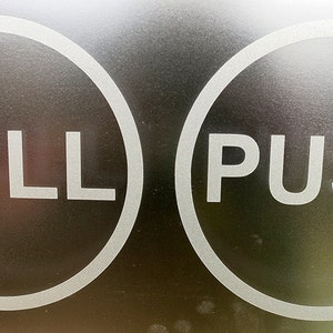 Könnte beinhalten: Nahaufnahme einer Tür mit zwei weißen kreisförmigen Schildern. Das linke Schild zeigt "PULL" und das rechte Schild "PUSH". Die Schilder befinden sich auf einer dunklen, möglicherweise metallischen Oberfläche.