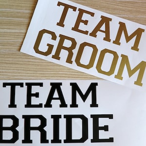 Peut inclure: Deux pancartes avec le texte "Team Groom" en lettres dorées et "Team Bride" en lettres noires. Les pancartes sont sur une surface en bois.