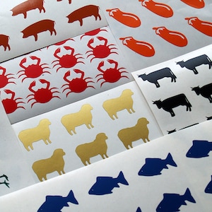 Op de afbeelding: Een vel stickers met verschillende dierenontwerpen. De stickers zijn gedrukt op wit papier en bevatten rode krabben, gouden schapen, zwarte koeien, blauwe vissen en bruine varkens.