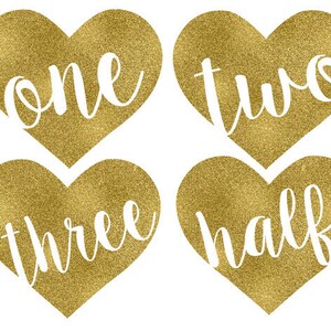 May include: Four gold glitter heart-shaped decorations with the words "one", "two", "three", and "half" written in white.