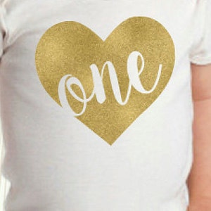 May include: White baby onesie with a gold glitter heart that says "one" in white cursive lettering.