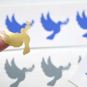 May include: A gold-colored paper dove with an olive branch in its beak. The dove is on a finger, and there are several blue and gray paper doves in the background.