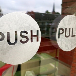 Può includere: Due adesivi rotondi bianchi su una porta di vetro. L'adesivo a sinistra dice "PUSH" e l'adesivo a destra dice "PULL".