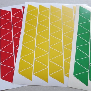 Puede incluir: Tres hojas de papel con filas de triángulos blancos sobre un fondo blanco. Los triángulos están rellenos de rojo, amarillo y verde.