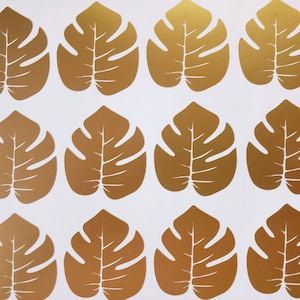 Könnte beinhalten: Zwölf goldene Monstera-Blatt-Aufkleber aus Folie. Die Blätter sind dunkelgoldfarben und haben eine glänzende Oberfläche.