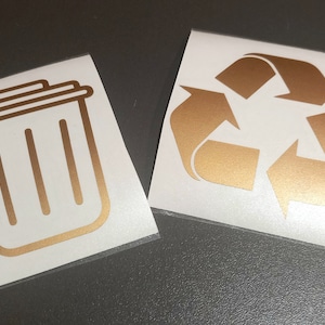Puede incluir: Dos pegatinas de vinilo doradas sobre fondos blancos. Una pegatina es un icono de cubo de basura. La otra pegatina es un símbolo de reciclaje.