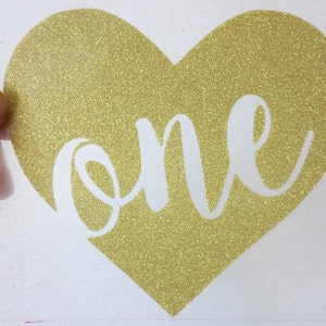 May include: Gold glitter heart-shaped sticker with the word "one" written in white cursive lettering.