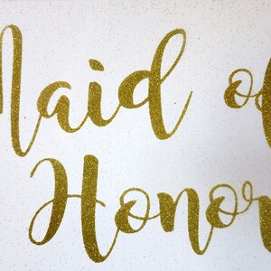 Könnte beinhalten: Goldene Glitzerbuchstaben auf weißem Hintergrund mit der Aufschrift "Maid of Honor".