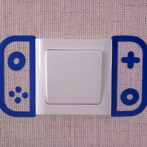 May include: A white light switch with a blue outline of a game controller on each side. The controller outlines feature button and directional pad designs. The switch is mounted on a textured, light-colored wall.