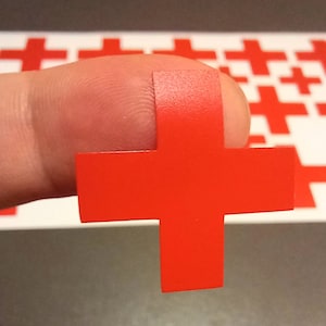 Może przedstawiać: Czerwona naklejka w kształcie krzyża, trzymana palcem, na tle wielu innych czerwonych naklejek w kształcie krzyża.
