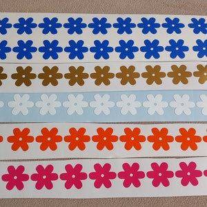 May include: Five rows of colorful flower-shaped stickers. The top row is blue, the second row is gold, the third row is white, the fourth row is orange, and the bottom row is pink.