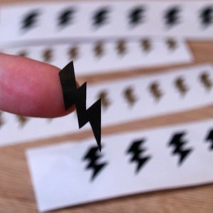 May include: A black lightning bolt sticker on a finger, with several other lightning bolt stickers on white paper in the background.