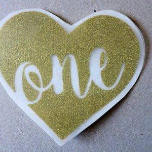 May include: Gold glitter heart-shaped sticker with the word "one" cut out in the center.