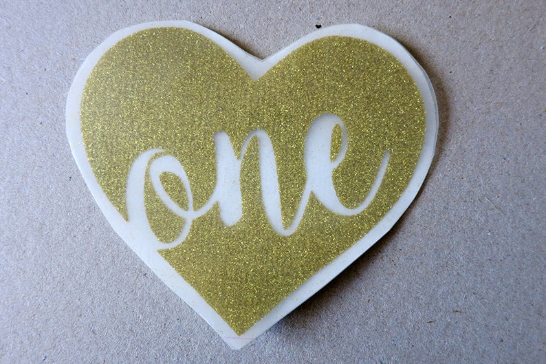 May include: Gold glitter heart-shaped sticker with the word "one" cut out in the center.