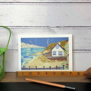 May include: A greeting card featuring a coastal scene with a white house, blue windows, and a blue sky. The card is displayed with a green lantern, a wooden fish, a ruler, and a pencil.