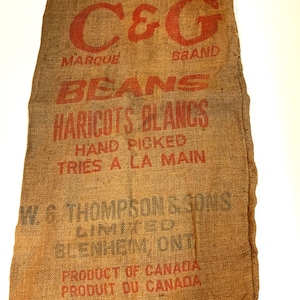 以下が含まれることがあります： 赤と青の印刷されたテキストが書かれた茶色のジュートの袋。テキストには「C&G Marque Brand Beans Haricots Blancs Hand Picked Tries a la Main W.G Thompson & Sons Limited Blenheim, Ont Product of Canada Produit du Canada」と書いてあります。