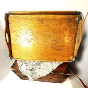 May include: A rectangular wooden serving tray featuring a natural light brown wood grain finish, integrated side handles, and polished chrome metal trim along the top edges. The tray has a flat base and curved corners, showcasing a smooth surface.