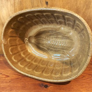 May include: A vintage brown ceramic mould shaped like an ear of corn. The mould has a textured surface and is designed for baking cornbread.